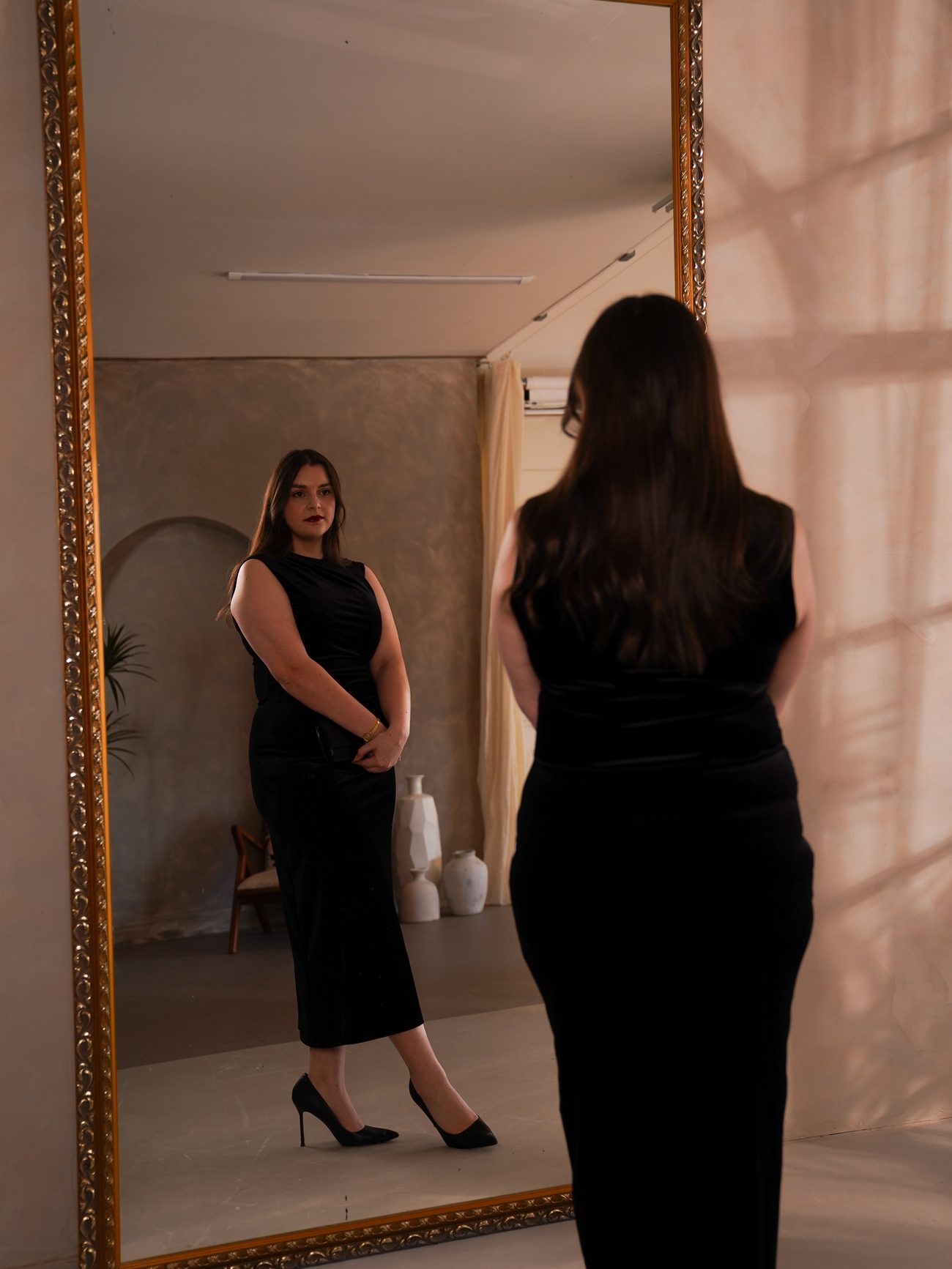 Frederike Behrens steht in einem schwarzen Samtkleid vor einem großen Standspiegel und betrachtet ihr Spiegelbild.