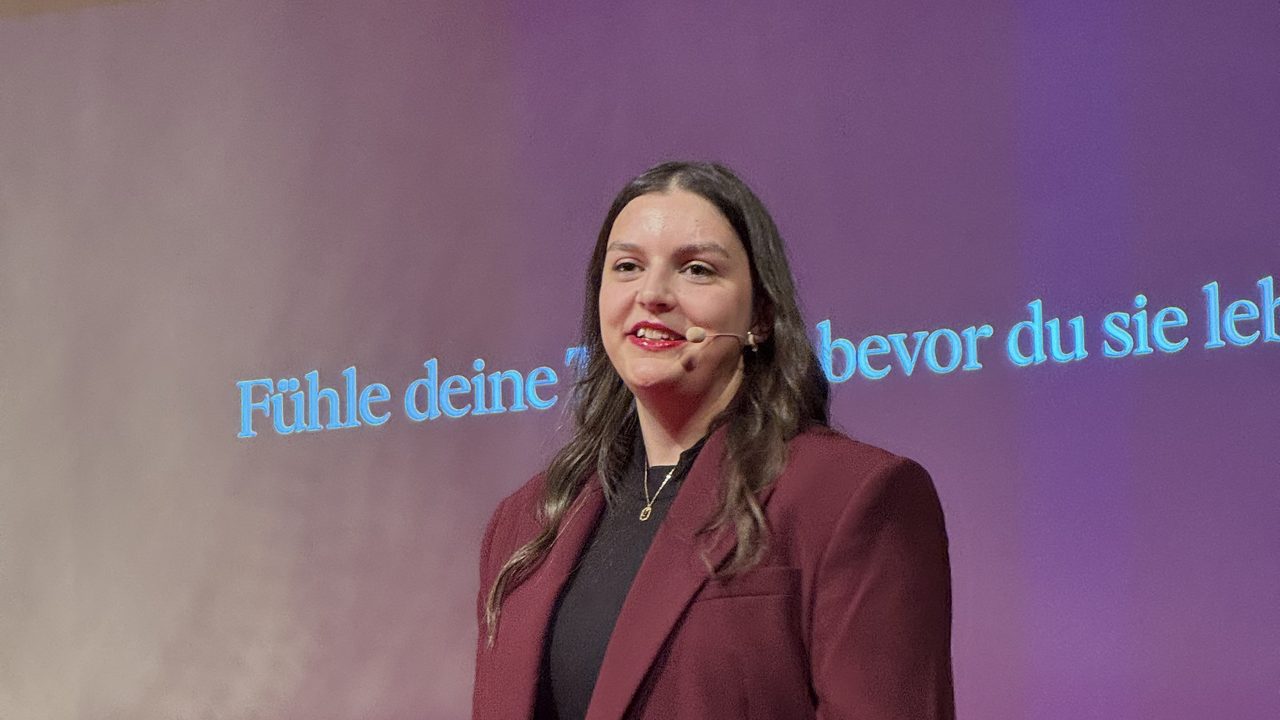 Frederike Behrens auf der B&uuml;hne mit Headset-Mikrofon vor einer gro&szlig;en Projektionsfl&auml;che.